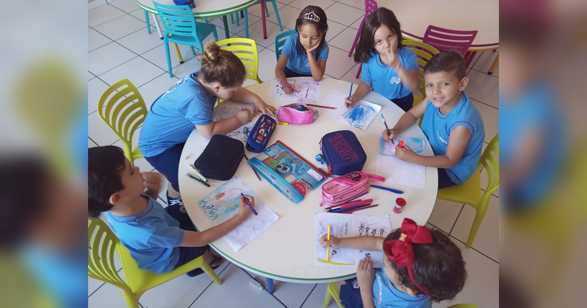 Seis pessoas, cinco crianças e uma adulta, estão sentadas em torno de uma mesa redonda branca com borda verde, em uma sala de aula. Tema - psicopedagogia infantil na Escola Aclive, Setor Vila Rosa, Goiânia, Goiás.
