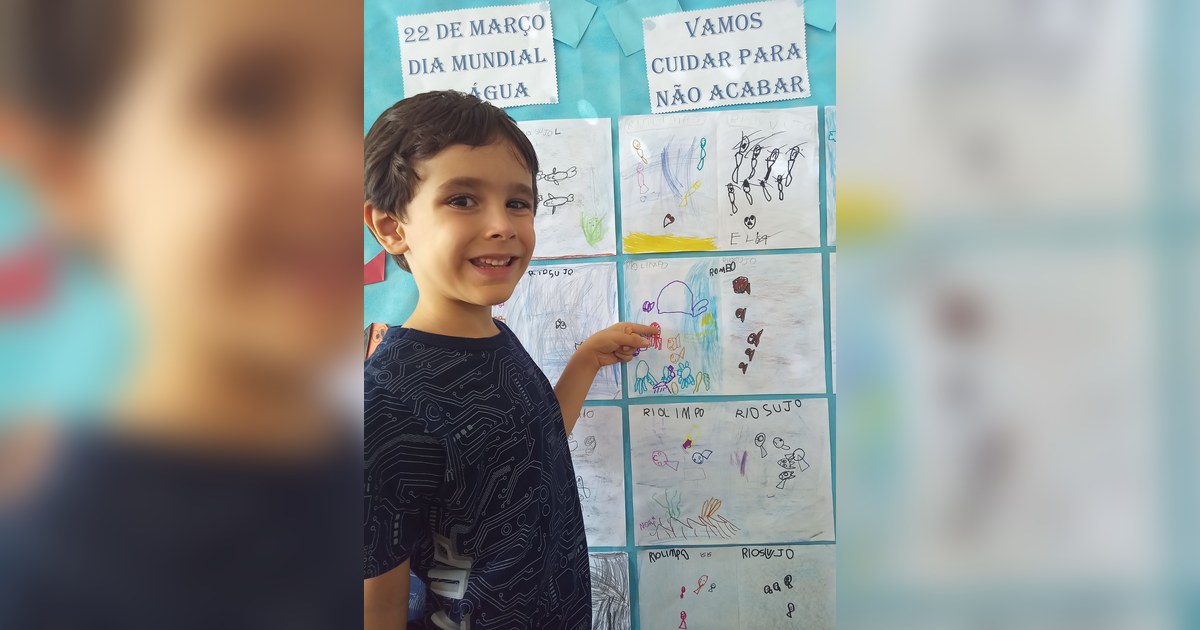 Uma criança, um menino de cabelo castanho, sorri para a câmera enquanto aponta com o dedo indicador da mão direita para um mural de trabalhos infantis. Tema - educação ambiental infantil na Escola Aclive, Setor Vila Rosa, Goiânia, Goiás.