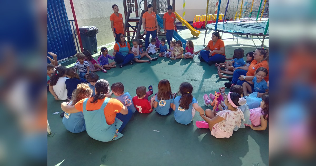 um vibrante momento de roda de conversa ao ar livre na Escola Infantil e Berçário Aclive, onde crianças de diversas idades interagem alegremente com as educadoras em um ambiente acolhedor e estimulante. Tema - educação socioemocional infantil na Escola Aclive, Setor Vila Rosa, Goi
