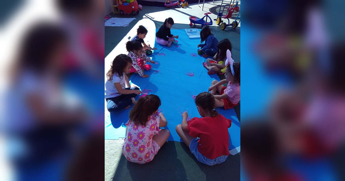 As crianças estão sentadas no chão de um pátio externo, formando um círculo ao redor de uma grande lona azul. Tema - atividades ao ar livre educação infantil na Escola Aclive, Setor Vila Rosa, Goiânia, Goiás.