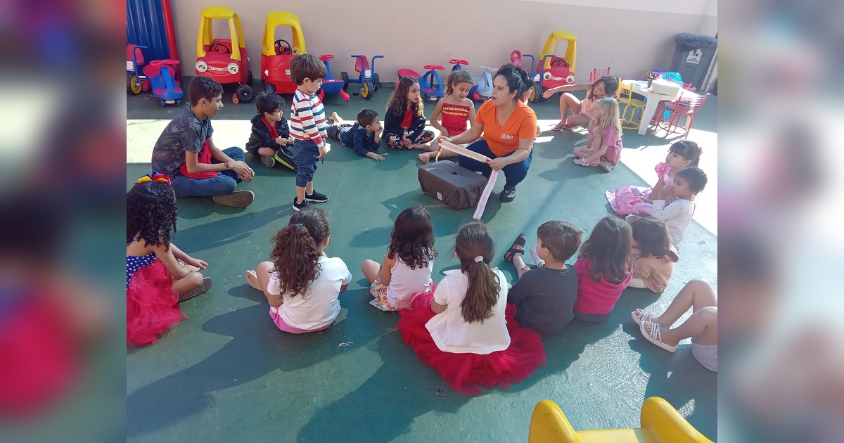 A foto exibe um grupo de aproximadamente vinte crianças e um adolescente/jovem, sentados ou agachados em semicírculo no chão, observando uma mulher adulta (provavelmente educadora) que está agachada no centro. Tema - preparar criança para escola na Escola Aclive, Setor Vila Rosa,
