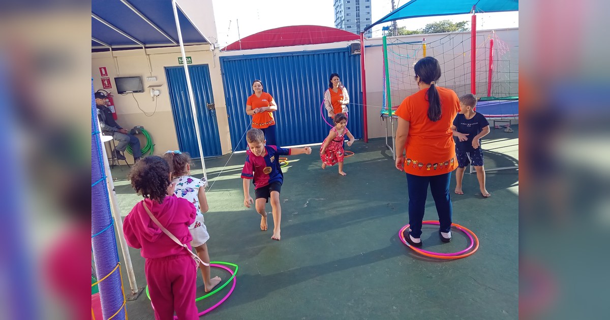 Sete crianças e três adultos estão visíveis em um pátio externo de uma escola infantil. Tema - coordenação motora ampla na Escola Aclive, Setor Vila Rosa, Goiânia, Goiás.
