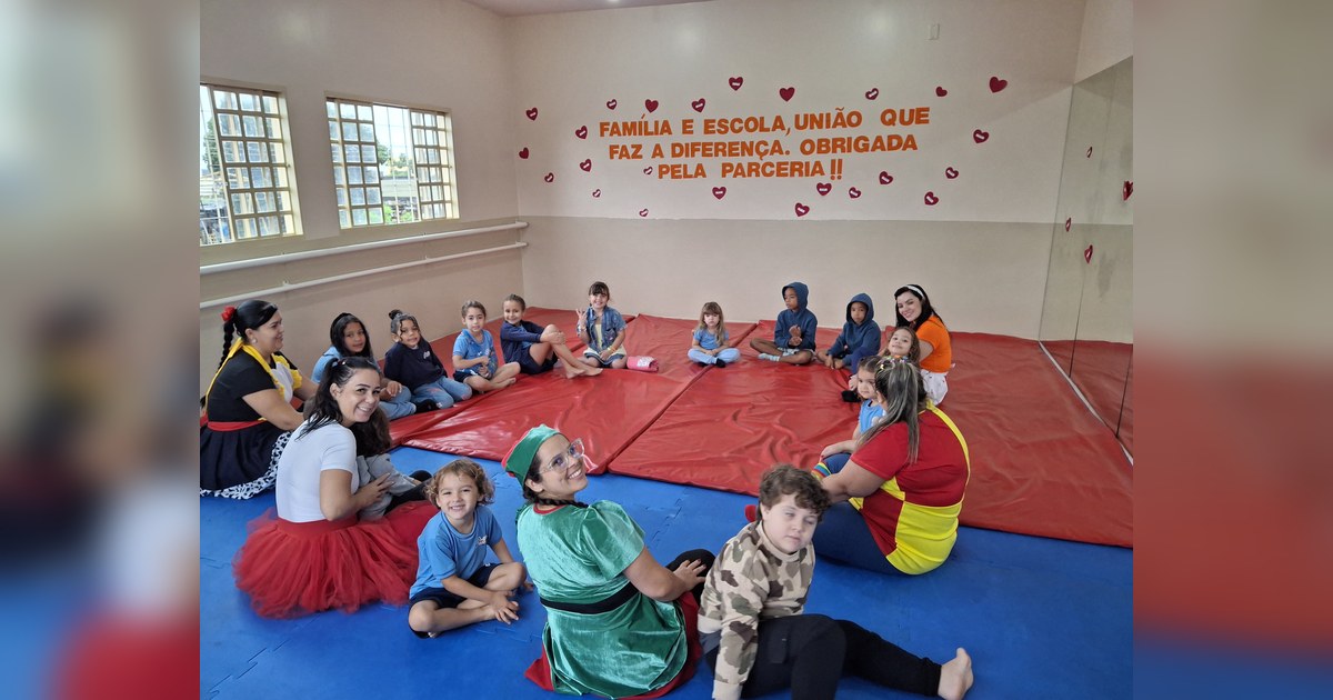Na foto, quinze crianças e quatro mulheres adultas estão sentadas em um ambiente interno espaçoso. Tema - leitura na educação infantil na Escola Aclive, Setor Vila Rosa, Goiânia, Goiás.