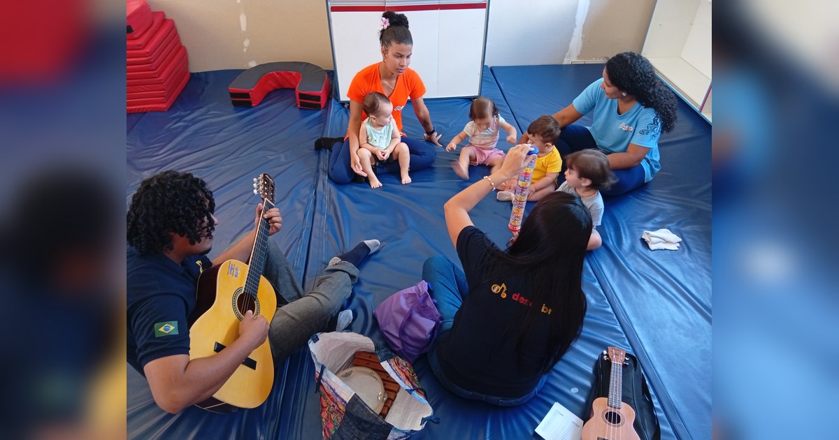 Seis adultos e cinco bebês ou crianças pequenas estão sentados no chão coberto por tatames azuis escuros em um ambiente interno. Tema - desenvolvimento infantil na Escola Aclive, Setor Vila Rosa, Goiânia, Goiás.
