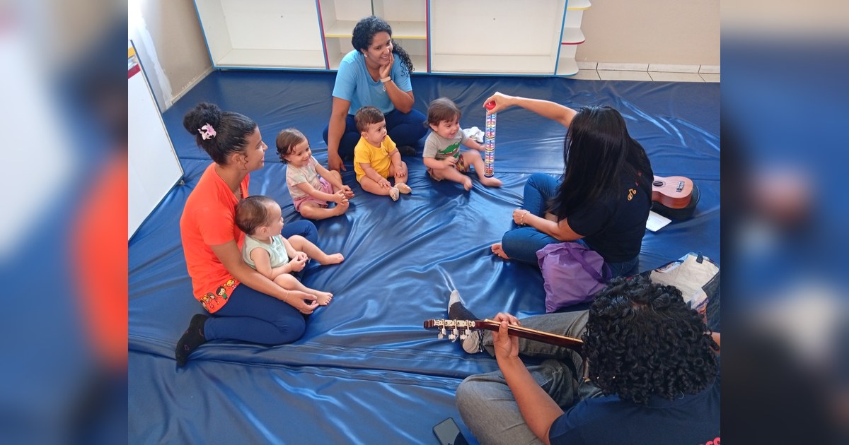 Quatro adultas e quatro bebês/crianças pequenas estão sentados no chão, sobre um grande tapete acolchoado azul escuro. Tema - rotina educação infantil na Escola Aclive, Setor Vila Rosa, Goiânia, Goiás.
