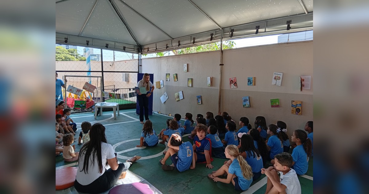 Três mulheres adultas e aproximadamente trinta crianças, variando de bebês a pré-escolares, estão reunidas em um espaço coberto. Tema - contação de histórias educação infantil na Escola Aclive, Setor Vila Rosa, Goiânia, Goiás.
